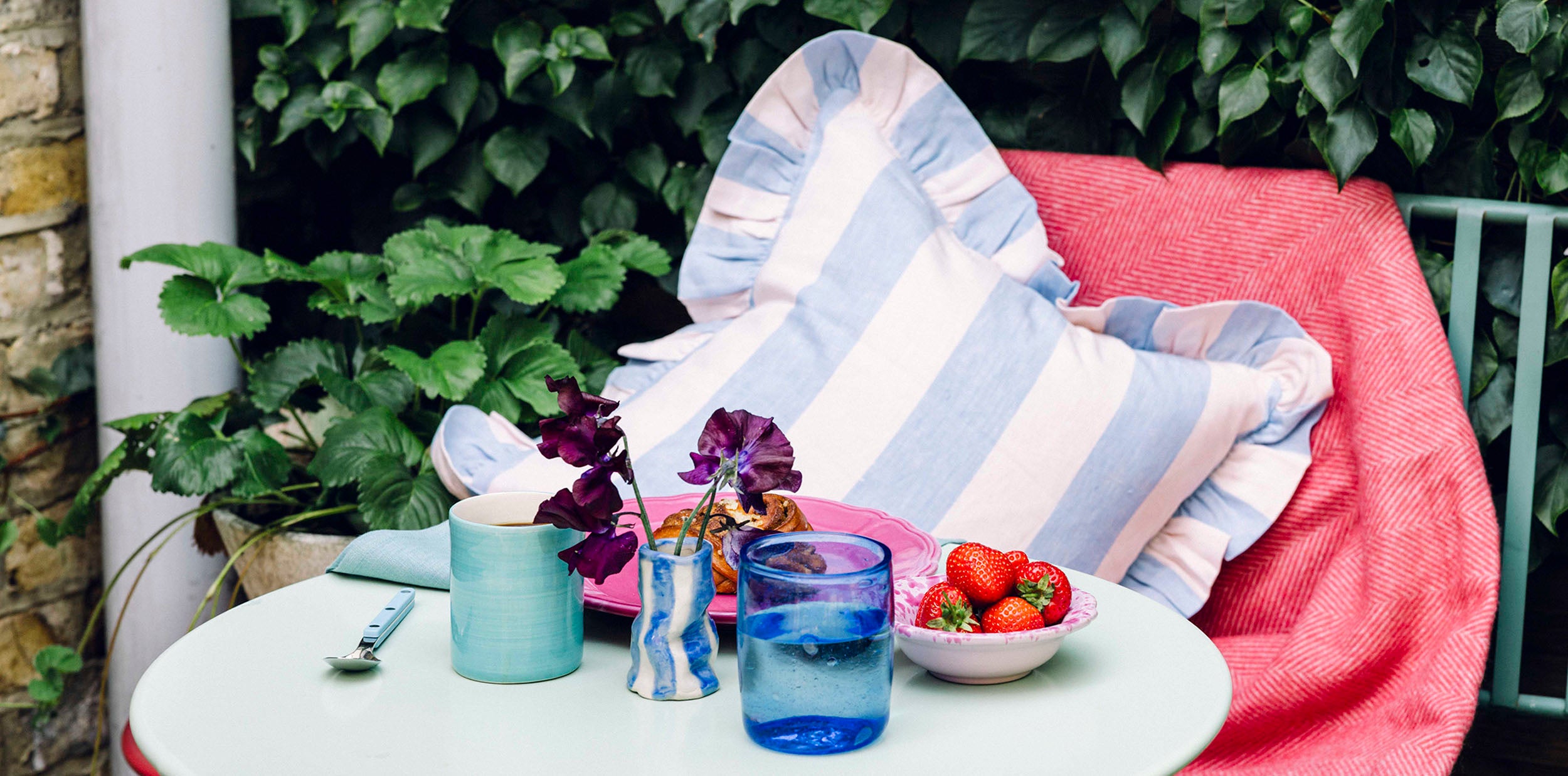 Garden table with a blue glass and mug and pink plate and blue striped frill cushion